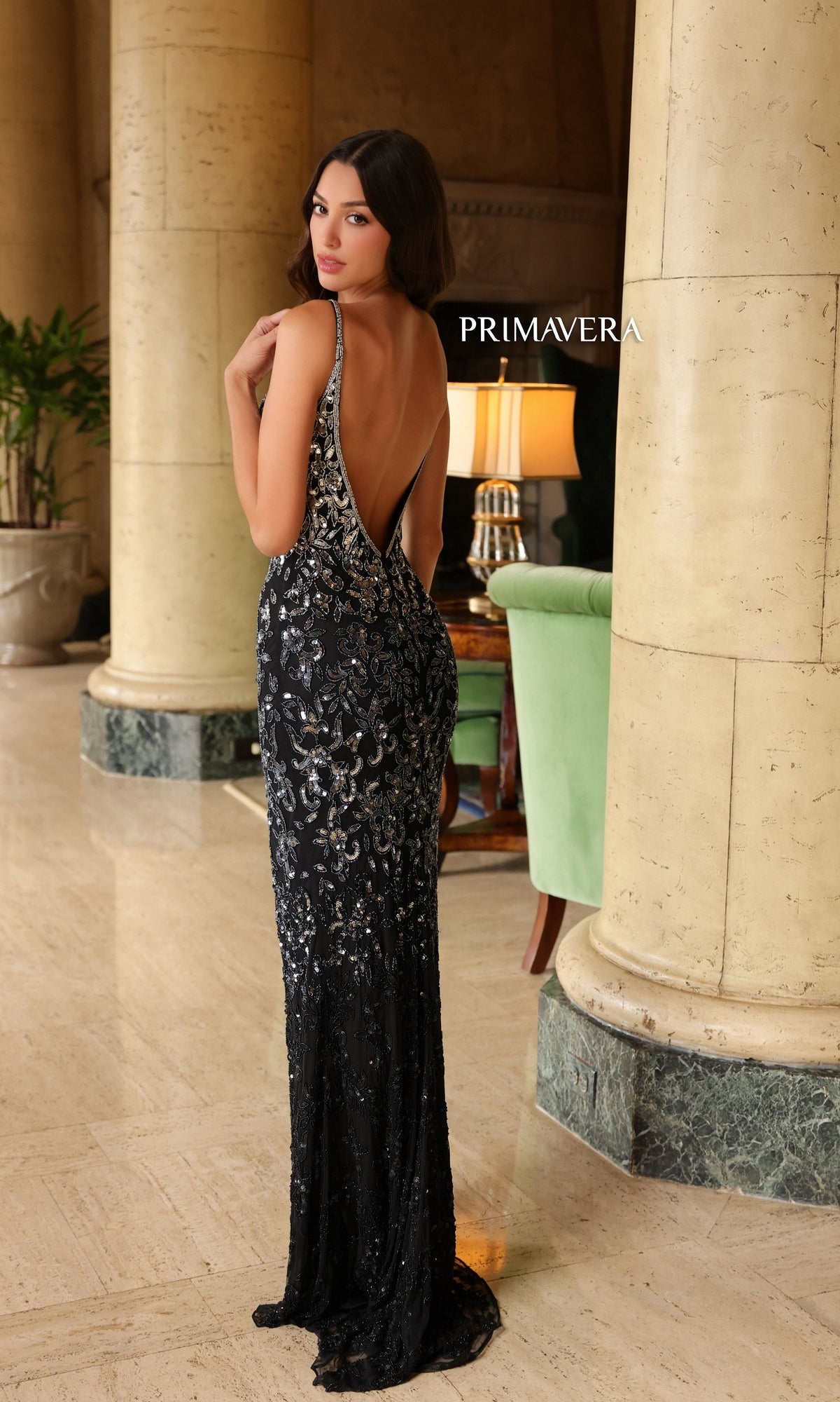 A woman stands indoors wearing the Primavera Ombre-Beaded Long Formal Gown 4150, a black, floor-length sequin dress with a low back. She looks over her shoulder with elegant columns, a lamp, and a green chair in the background.